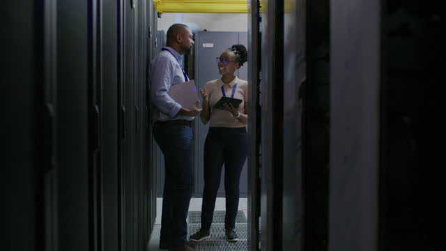 Man, woman and talking with tablet in server room, IT and computer programming. Cybersecurity team, digital technology and data center for coding backup, engineering and planning maintenance update