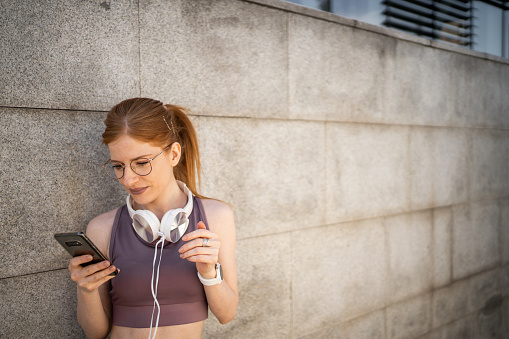 Young woman with orange hair, redhead, young athlete resting after training leaning against concrete wall, looking at phone and wearing headphones