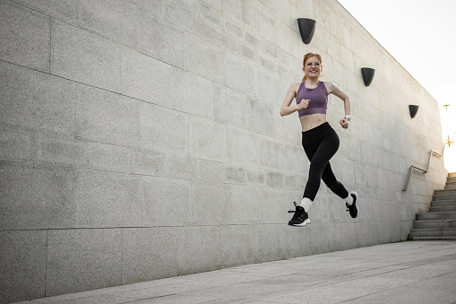 Young red-haired fitness girl runs and jumps next to the concrete wall, takes care of her physical strength and is happy to train