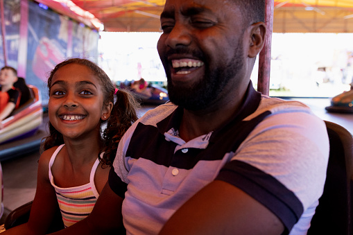 A close-up of a man and his young daughter enjoying a day out at the fair in Newcastle upon Tyne, North East England. They are on the bumper cars and laughing together, the young girl is looking at the camera.