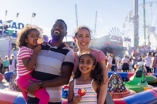 A family enjoying a day out at the fair in Newcastle upon Tyne, North East England. They are standing together, looking at the camera and smiling while the two young girls eat toffee apples with fairground rides behind them. The father is carrying the youngest daughter.