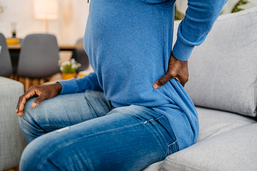 Close-up of tired young handsome black man having back pain while sitting on the sofa at home.