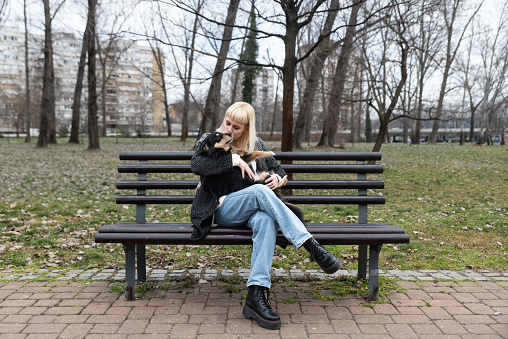 Young beautiful hipster woman sitting on a bench in the park with her adopted dog that saved his life from the street and the animal shelter and keeps him in her arms, gives him love food and care
