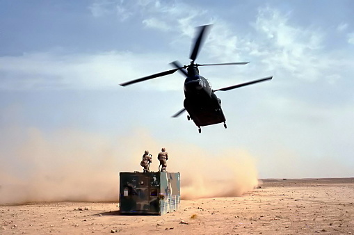 A US Army Chinook helicopter from the 101st Airborne Division swoops in over the Saudi Arabian desert while two soldiers with clevis hooks are positioned to attach them to the fore  and aft cargo hooks on the aircraft during the air war phase of the first Gulf War in 1991.