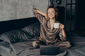 Relaxed young brunette female in satin pajama having her cup of coffee in bed and checking emails on laptop