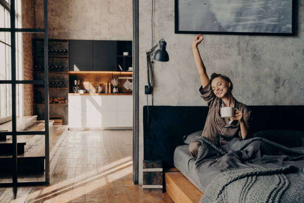 Young beautiful woman sitting on bed stretching with her hand up while holding cup of coffee Energy drink. Young beautiful positive european woman sitting on bed stretching with her hand up while holding cup of coffee with another hand, trying to wake up before going to shower in morning woman hot shower stock pictures, royalty-free photos & images