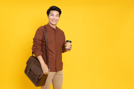 Happy young Asian man standing with leather bag and hot coffee cup isolated on yellow background