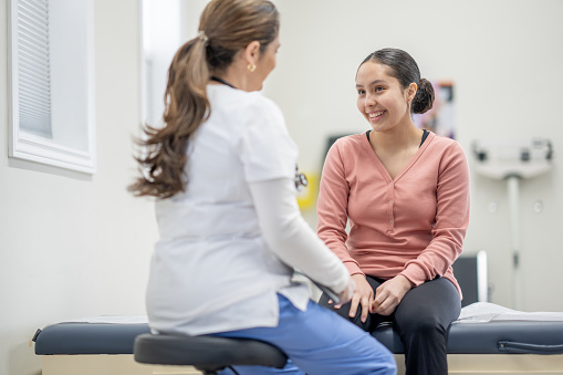 A young female teenager sits up on an exam table at the doctors during a routine check-up.  She is dressed casually and her female nurse of Hispanic decent, is seated in front of her as they talk.