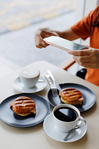 A young woman is using her smartphone to capture an image of a cafe table set with coffee cups and toasted bread