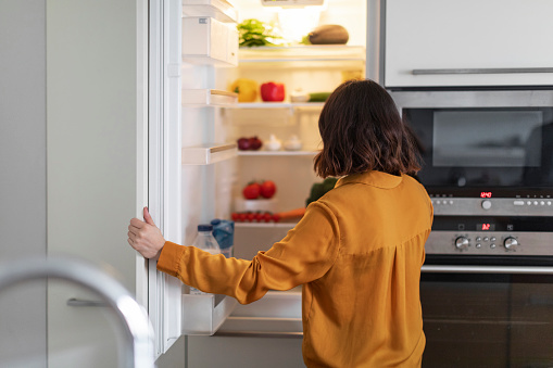 Unrecognizable Young Woman Opening Fridge In Kitchen And Looking Inside, Hungry Brunette Lady Standing Near Open Refrigerator At Home, Starving Female Checking Food Or Drinks, Free Space