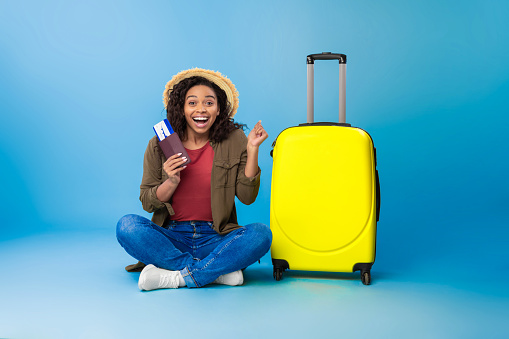 Excited young black woman with bright suitcase, passport and plane ticket sitting cross legged and making YES gesture on blue studio background. Summer vacation, tourism concept