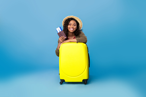 Happy black lady in casual wear sitting behind bright suitcase, holding passport and flight tickets, going on summer vacation on blue studio background. Tourism, trip, travelling concept