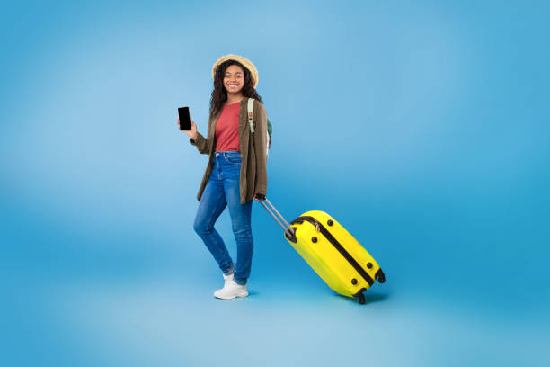 istock Cheery young black woman with yellow suitcase holding smartphone with empty screen, offering mockup for website 1479370233
