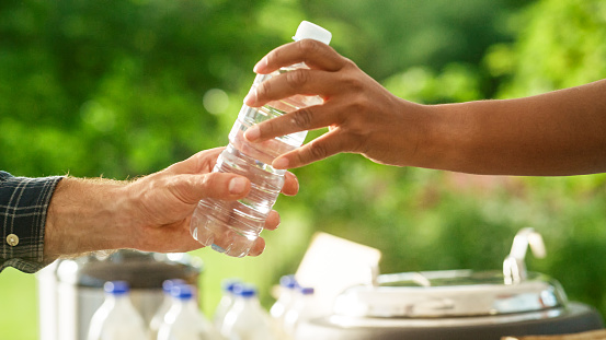 Close Up of a Anonymous Person Handing Over a Water Bottle to Another Person. Green Background in Nature. Outdoors Fourt Court Selling Drinks. Ecology, Healthcare and Hydration Concept.