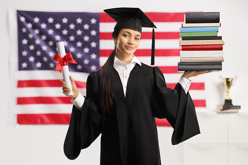 Female graduate student in a gown holding a pile of books and a diploma with USA flag in the background