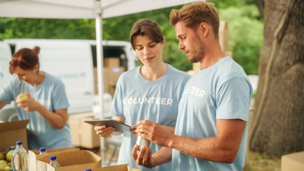 Team of Young Volunteers Helping in a Local Community Food Bank, Preparing Free Meal Rations to Low-Income People in a Park on a Sunny Day. Charity Workers Work in Humanitarian Aid Donation Center. stock photo