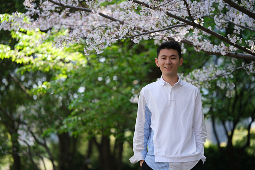 front portrait of smile handsome Asian young man in casual shirt, hands in pocket. Looking at camera under white sakura blossom tree