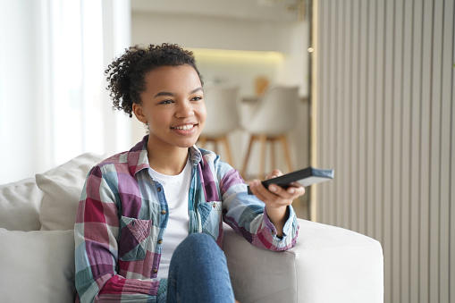 Attractive teenage girl is watching it and shifts channels with remote controller. Reality show or series watching. Positive african american or hispanic young woman has weekend leisure at home.
