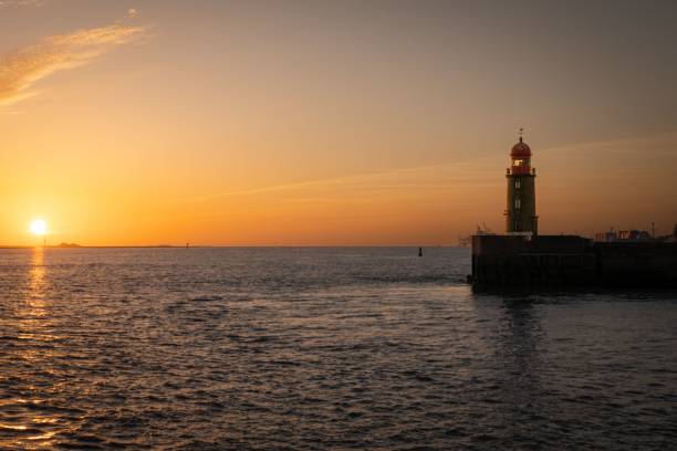 A picturesque lighthouse illuminated by the setting sun, which casts a few clouds in the sky