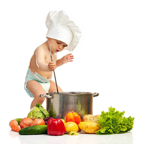 Portrait of one-year old boy in chef's hat with big ladle, casserole, and fresh vegetables over white background