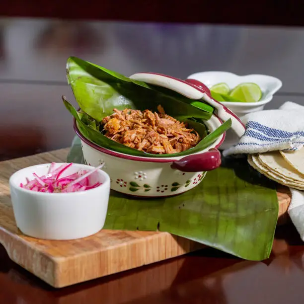 Photo of cochinita pibil placed on banana leaves accompanied with red onion, tortillas and lemons