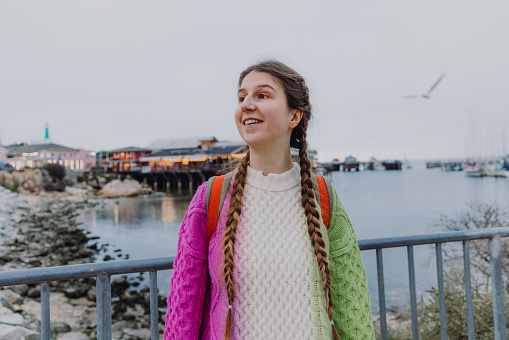 Happy female with long hair wearing colorful sweater walking on the pier at Old Fisherman's Wharf, contemplating old houses by the ocean in Monterey Peninsula, the USA