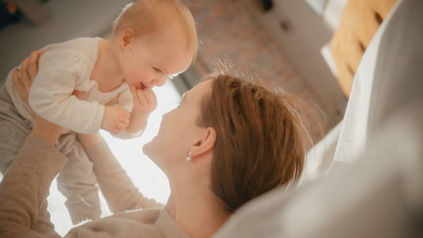 счастливая мама играет с милым новорожденным ребенком. мама связывается с малышом, держится над ней, целует младенца в щеку. понятие детств� - mother baby new kissing стоковые фото и изображения