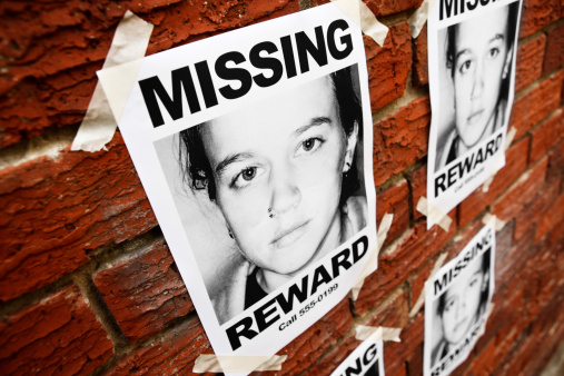 Looking down at a group of identical Missing posters of a sad young girl, taped to a brick wall, possibly inside a police station. 