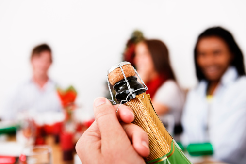 Hand opening champagne in foreground, Christmas-decorated table and tree in background with young people. Could be an office Christmas party or a gathering of friends at home. 