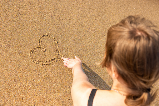 Woman drawing heart shape on the sand with finger at seaside on a sunny day - Top view of a caucasian blond woman in love - concept and symbol of love