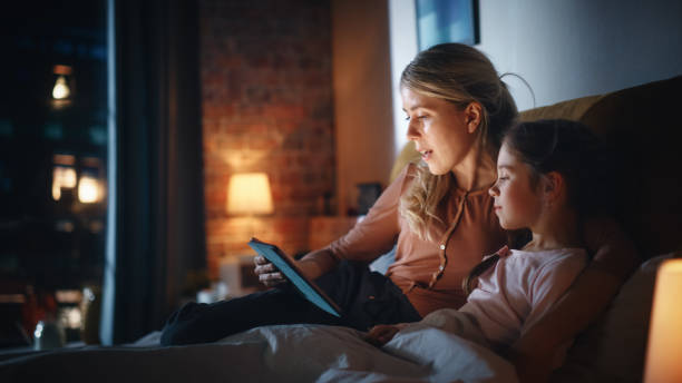 istock Mother Reading a Fairytale from a Tablet Computer to Her Lovely Daughter in Bed Before Going to Sleep. Young Babysitter Caring for Little Girl, Reading Online Stories in the Evening at Home. 1479420646