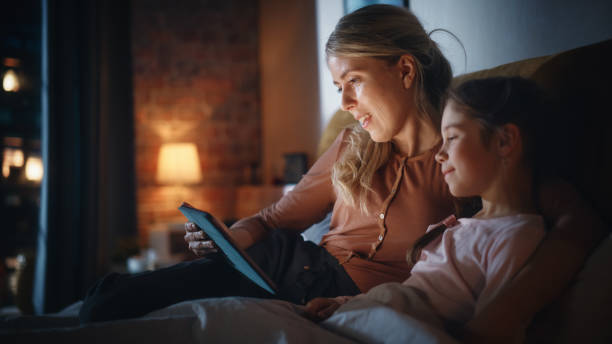 istock Mother Reading a Fairytale from a Tablet Computer to Her Lovely Daughter in Bed Before Going to Sleep. Young Babysitter Caring for Little Girl, Reading Online Stories in the Evening at Home. 1479420693