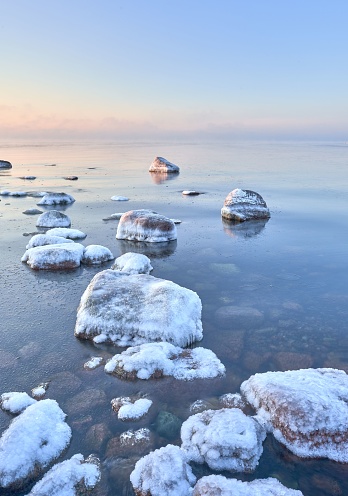 A tranquil shoreline in winter, with snow-covered rocks and a partially frozen surface of the sea