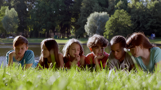 Joyful multiracial preteen friends lying on park grass together. Portrait of happy teen kids lying in row on green lawn outdoors and talking having fun together on school break