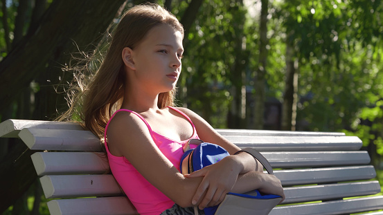 Sad lonely teenage girl sitting on bench in park. Upset preteen schoolgirl with backpack sit on bench outdoors depressed because of school or friends