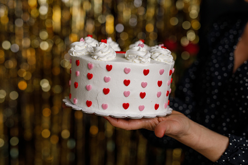 Close up shot of unrecognizable woman standing against a gold colored fringe curtain and holding a cute little cake decorated with pink and red colored love hearts.