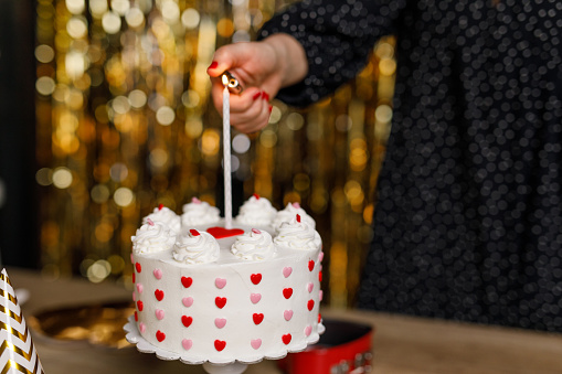 Close up shot of unrecognizable woman standing against a gold colored fringe curtain and lighting a birthday candle on a cute cake, decorated with pink and red colored love hearts, during a celebratory party.