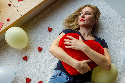 Directly above shot of happy mid adult woman, in love, lying on the floor, surrounded with love hearts and balloons, embracing a heart shaped pillow and, looking away, smiling and daydreaming about her loved one.