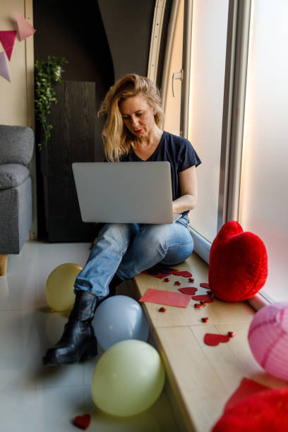 frau sitzt auf dem fensterbrett, benutzt einen laptop und träumt von ihrem liebsten - working at home calendar laptop women stock-fotos und bilder