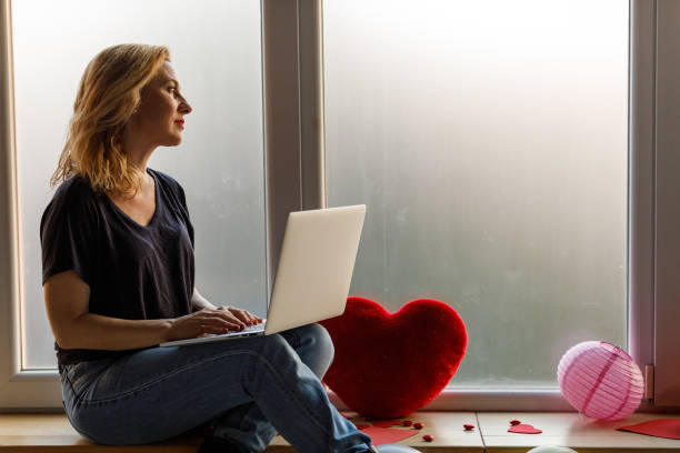 frau sitzt auf dem fensterbrett, benutzt einen laptop und träumt von ihrem liebsten - working at home calendar laptop women stock-fotos und bilder