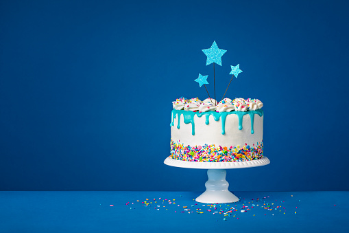 Fun White Birthday cake with trendy teal ganache drips, colorful sprinkles and star toppers over a dark blue background. Copy space.