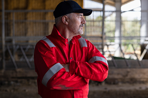 A waist-up portrait of a male farmer with grey hair and a beard, wearing red overalls and a cap. He is looking over his shoulder to the right of the frame with his arms crossed, and is standing in an agricultural building on a farm in Northumberland, North East England.