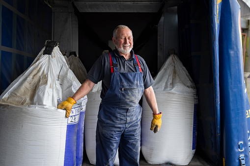 A shot of a male delivery person dropping off a delivery of nitrogen sulphur fertiliser to a sustainable farm in Northumberland, North East England. He is standing in the back of the delivery truck, smiling and wearing overalls and gloves, with one hand on a fertiliser sack.