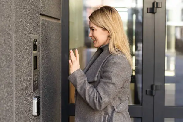 Happy woman ringing on doorbell at building entrance. Using intercom.