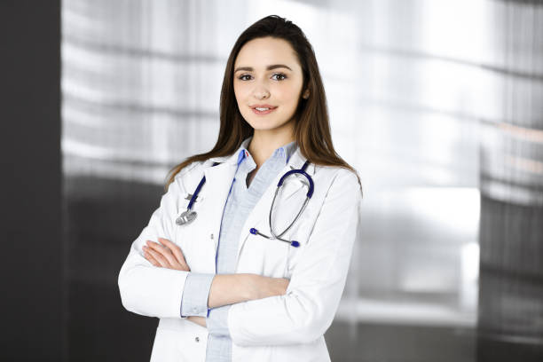 une jeune femme-médecin souriante se tient debout, les bras croisés, dans le bureau d’une clinique. portrait d’une femme médecin sympathique. service médical parfait dans un hôpital. concept de médecine - portrait doctor paramedic professional occupation photos et images de collection