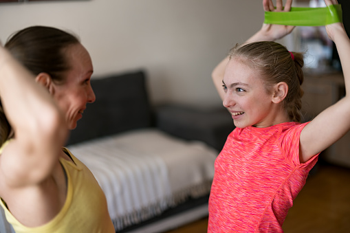 Mature mother and teenage daughter doing arms exercise with resistance band at home