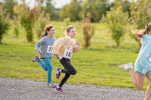 A multi-ethnic group of school aged children are seen running down a gravel trail during a running race.  They are each dressed comfortably and are wearing numbered bibs.