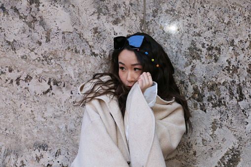 A Chinese woman showing shyness in front of a stone wall. She is wearing long, black tousled hair, sunglasses on top of her head, and a long sleeved white dress.