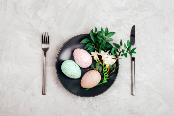 istock Painted Easter eggs and freesia white flower on black plate on gray textured background. Holiday table setting. Top view, flat lay 1479460015