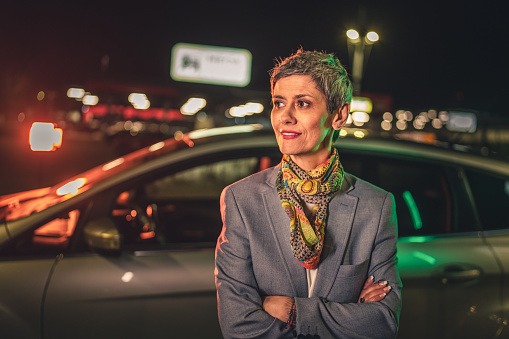 portrait of one senior woman modern mature caucasian female with gray short hair stand in the city at night in front of the car real person copy space waist up